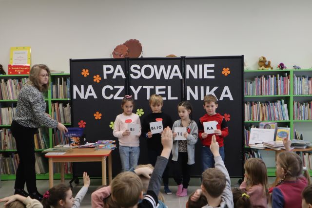 dzieci siedzą na zielonym dywanie, przed nimi stoją dzieci i trzymają w dłoniach katki z napisem biblioteka, w tle regały z książkami i czarne tablica