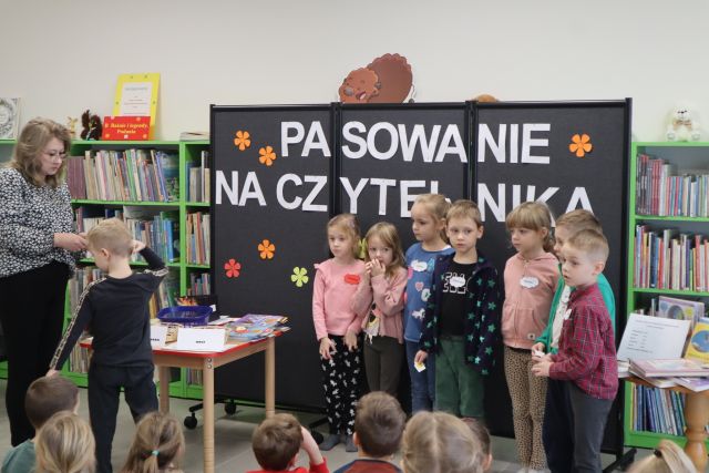 dzieci stoją przed czarną tablicą, w tle regały z książkami