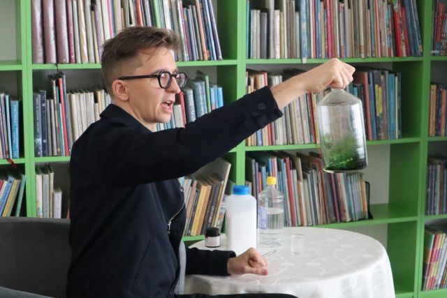 mężczyzna w czarnej marynarce siedzi na siwym fotelu i trzyma w ręku butelkę z kolorowym płynem, w tle regały z książkami