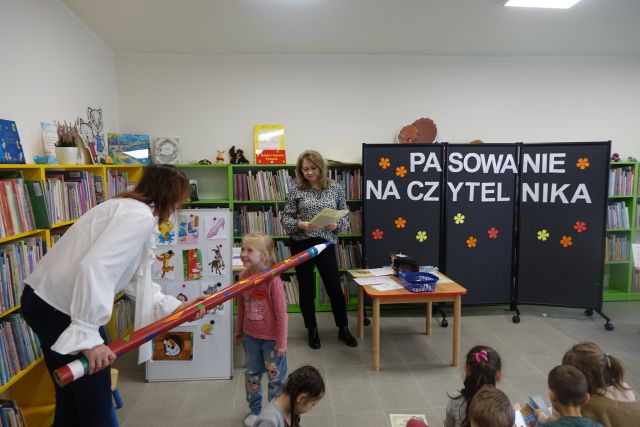 dziewczynka w różowej bluzce jest dużym czerwonym ołówkiem pasowana na czytelnika przez kobietę w białęj bluzce, w tle czarna tablica i regały z książkami