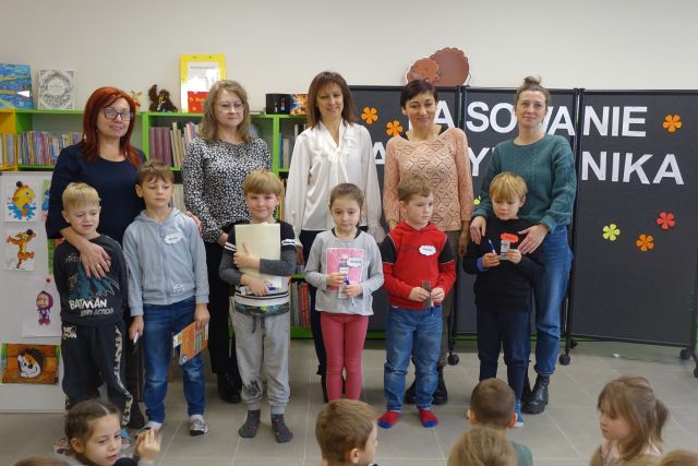 6 dzieci stoi, za nimi cztery dorosłe kobiety, w tle regały z książkami
