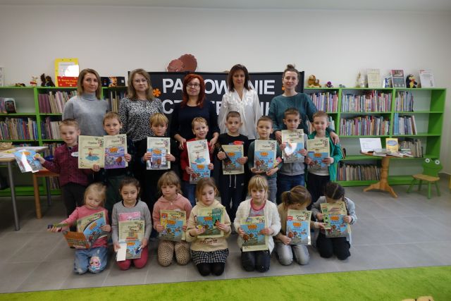w pierwszym rzędzie klęczą dzieci, za nimi stoją dzieci, a za nimi kobiety, w tle regały z książkami