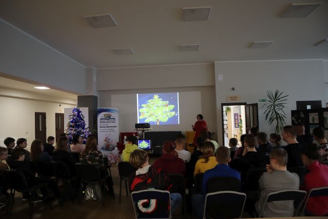uczestnicy spotkania siedzą na krzesłach, przed nimi stoi kobieta w czerwonej długiej bluzce, na ścianie wyświetlona prezentacja, z lewej strony ubrana choinka i plakat z informacją o dofinansowaniu