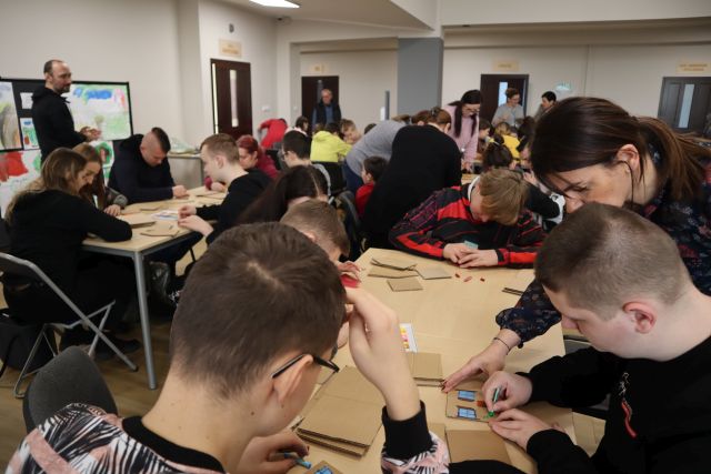uczestnicy warsztatów siedzą przy stolikach i kolorują domki z kartonów