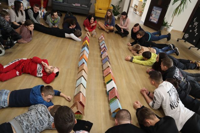 na podłodze leżą uczestnicy warsztatów, przed nimi ulica zbudowana z domków z kartonów