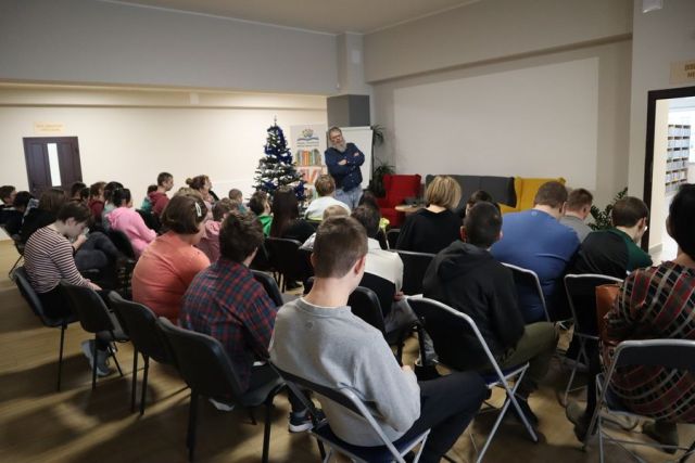 uczestnicy warsztatów siedzą na krzesłąch, przed nimi autor w siwym swetrze i jasnych dżinsach opowiada