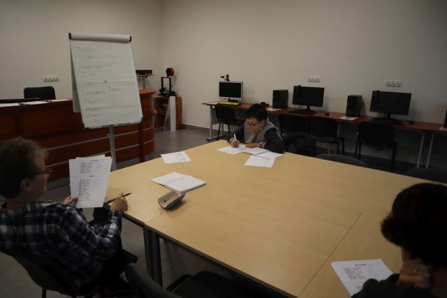 przy brązowych połączonych stołach siedzą trzy kobiety i piszą na kartkach, przed nimi flipchart