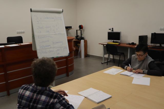 dwie kobiety siedzą przy brązowych połączonych stołach, przed nimi flipchart