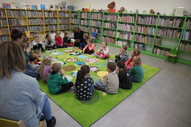 dzieci siedzą na zielonym dywanie, w tle regały z książkami