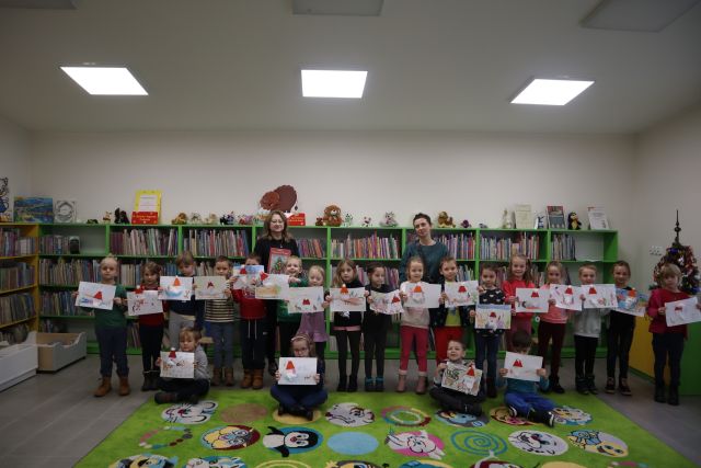 dzieci w pierwszym rzędzie kucają na zielonym dywanie, w drugim rzędzie stoją, wszyscy trzymają w dłoniach ilustarcje z mikołajami