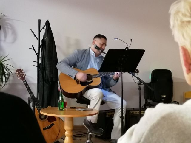 mężczyzna w siwej marynarce siedzi na wysokim krześle i gra na gitarze