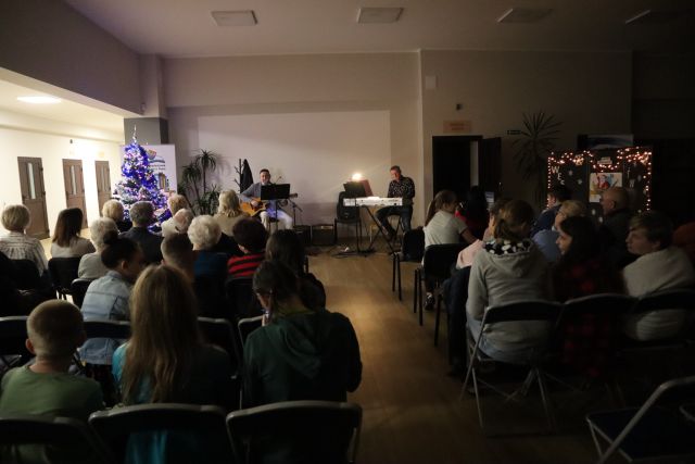 uczestnicy koncertu siedzą na krzesłach, przed nimi zespół muzyczny