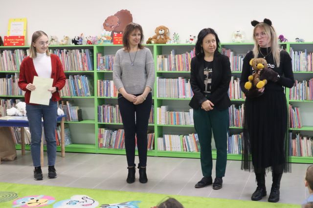 kobieta w siwej bluzce i granatowych spodniach, kobieta w czarnej marynarce i kobieta w stroju misia stoją przed dziećmi, w tle regały z książkami