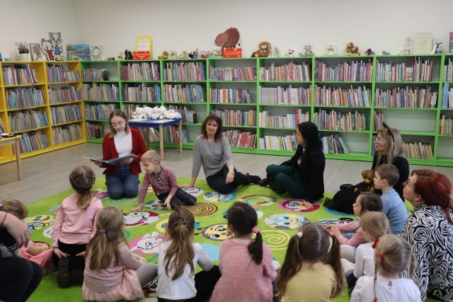 na zielonym dywanie siedzą przedszkolaki i 4 osoby dorosłe, w tle regały z książkami