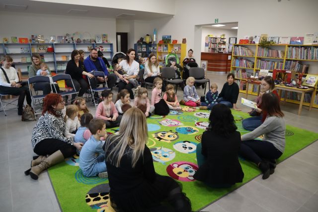 dzieci i dorosłe osoby siedzą na zielonym dywanie, w tle regały z książkami i część dorosłych osób i dzieci niepełnosprawnych siedzi na krzesłąch i wózkach inwalidzkich