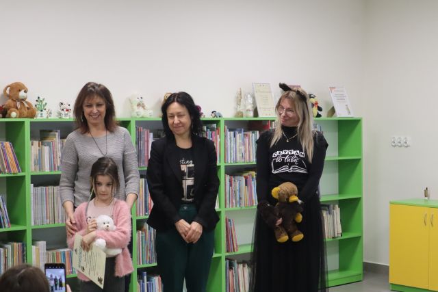 trzy kobiety stoją, przed nimi dziewczynka z misiem, w tle regały z książkami
