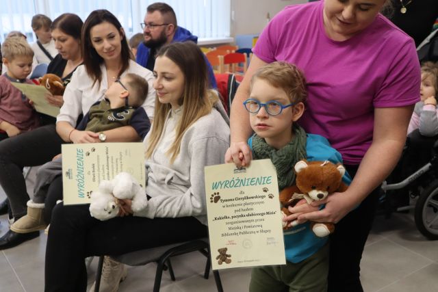 chłopiec w okularach na wózku inwalidzkim trzyma dyplom i misia, obok siedzą inni laureaci konkursu i ich opiekunowie