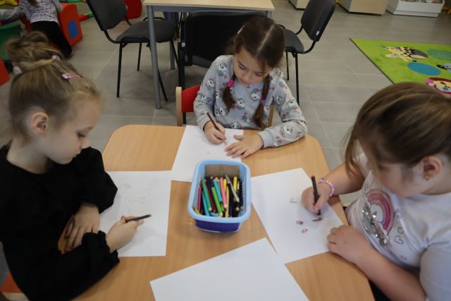 dzieci siedzą przy brązowym stole i malują na kartkach