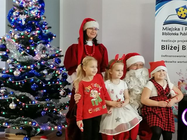 mikołąj siedzi na czerwonym fotelu, na jego kolanach siedzą dwie dziewczynki, za nimi stoi mikołąjowa, z lewej strony ubrana choinka