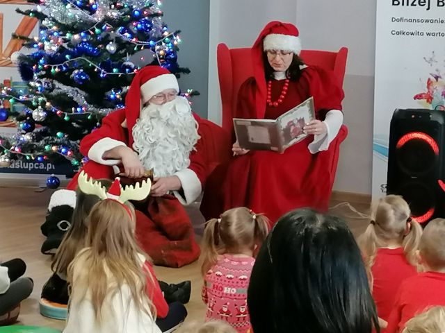 Mikołąjowa w czerwonej sukience i czapce siedzi na czerwonym fotelu i czyta książkę, Mikołaj siedzi na podłodze, w tle choinka
