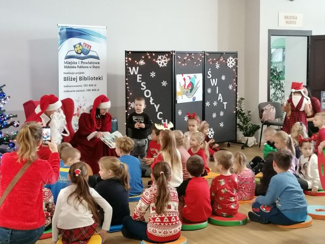 dzieci siedzą na podłodze, przed nimi na fotelu siedzi mikołajowa i czyta książkę, obok na podłodze siedzi mikołaj, w tle choinka