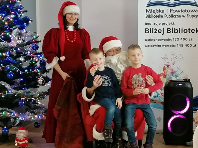 mikołąj siedzi na czerwonym fotelu, na jego kolanach siedzą dwaj chłopcy, obok stoi mikołąjowa