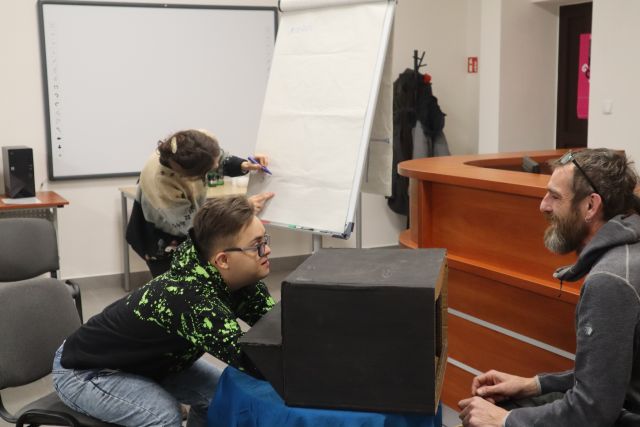 chłopiec maca w zakrytym siwym materiałęm kartonie nieznany przedmiot, przed nim siedzi mężczyzna w siwej bluzie z włosami w dredach, w tle brązowe biurko