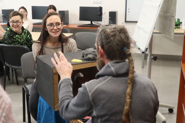 kobieta w siwej bluzce wyszukuje w kartonie zasłoniętym siwym materiałem nieznany przedmiot, przed nią siedzi tyłęm mężczyzna w siwej bluzie i z włąsami w dredach