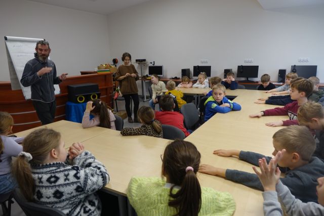 przy brązowych stołąch siedzą dzieci, przed nimi prowadzący warsztaty a w tle brązowe biurko