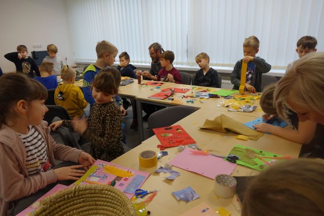 dzieci siedzą przy brązowych stołach i przyklejają różne kawałki materiałów na kartki