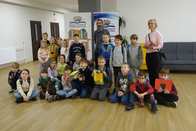 dzieci w pierwszym rzędzie kucają i trzymają w rękach wykonane przez siebie książki sensoryczne, za nimi stoją pozostali uczniowie i mężczyzna w siwej bluzie, w tle plakaty z nazwą projektu