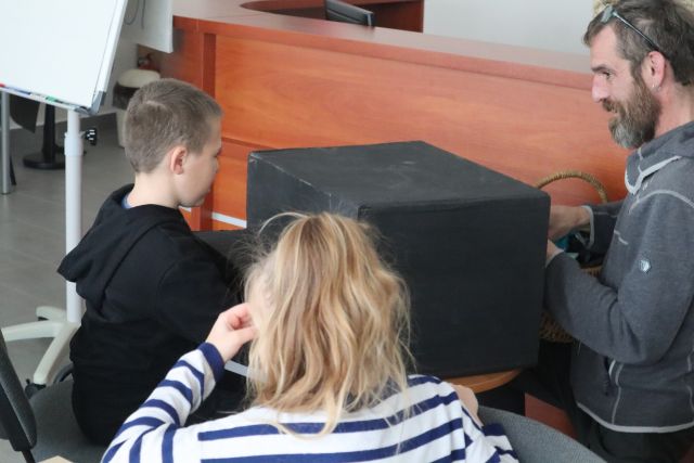 chłopiec w czarnej bluzie poszukuje przedmiotu w zakrytym siwym materiałęm kartonie, z drugiej strony mężczyzna w siwej bluzie i włosach dredach podaje ukryty przedmiot