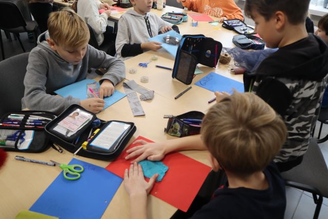 dzieci siedzą przy stołąch i wyklejają z różnych materiałów książki