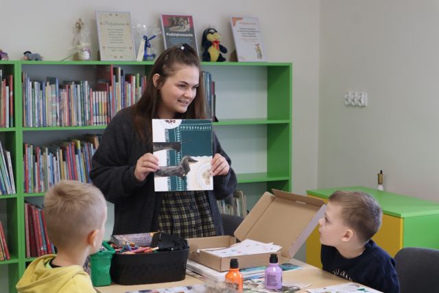 kobieta w ciemnym swetrze i spódnicy w biało-czarną kratkę pokazuje dzieciom ilustracje, w tle regały biblioteczne pokazuje dz