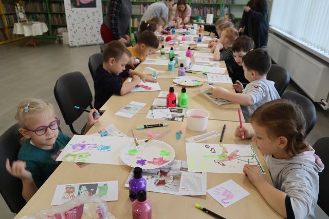dzieci siedzą przy brązowych stołach i malują farbami na kartkach