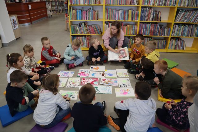 dzieci siedzą na podłodze, w środku mają namalowane przez siebie rysunki, kobieta klęczy przed nimi i pokazuje książkę, w tle żółte regały z książkami