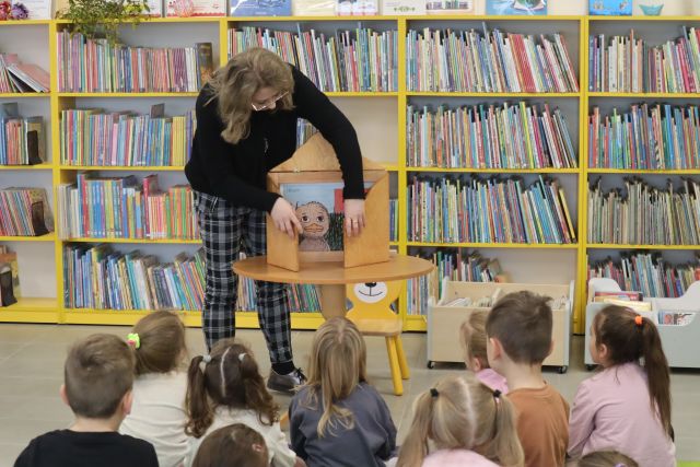 kobieta w jasnych włosach, czarnej bluzce i spodniach w kratkę otwiera teatrzyk kamishibai, przed nią na zielonym dywanie siedzą dzieci, w tle regały z książkami