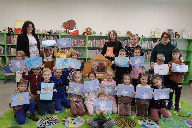 dzieci siedzą i stoją, w rękach trzymają wykonane przez siebie ilustracje łabędzi, w tle regały z książkami