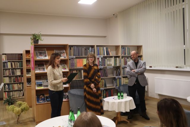 kobieta w kremowym swetrze czyta, obok stoi kobieta w sukience w żółto czarne pasy, obok niej za stolikiem z jasnym obrusem stoi mężczyzna w siwej marynarce, w tle regały z książkami