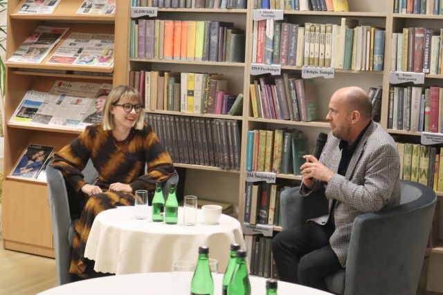 kobieta w sukience w żółto czarne pasy siedzi przy stoliku z jasnym obrusem, po drugiej stronie siedzi mężczyzna w siwej marynarce, w tle regały z książkami