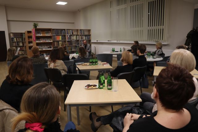 uczestnicy spotkania siedzą przy stolikach, w tle regały z książkami