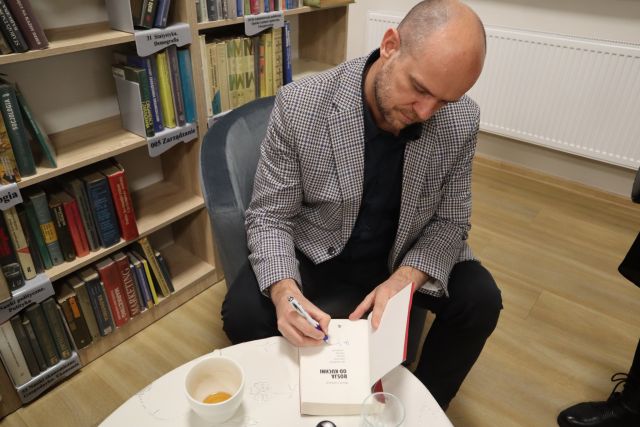 autor, mężczyzna w siwej marynarce siedzi na siwym fotelu i podpisuje książkę, w tle regały z książkami