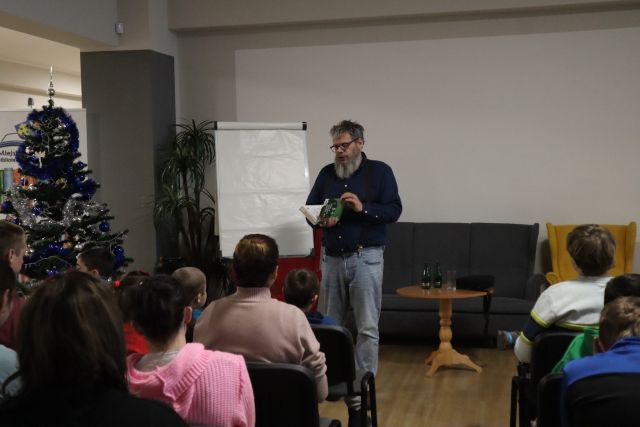uczestnicy spotkania siedzą na krzesłąch, przed nimi mężczyzna w ciemnym swetrze i jasnych dżinsach