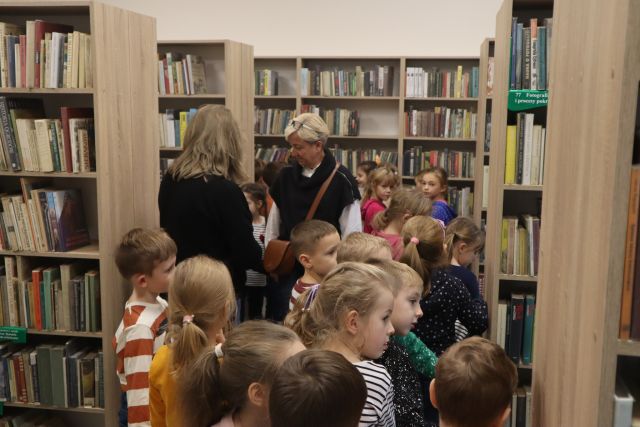 małe dzieci wśród regałów z książkami, przed nimi dwie kobiety w czarnych bluzkach