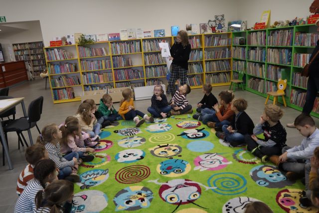 dzieci siedzą na zielonym dywanie, przed nimi stoi kobieta w czarnej bluzce i pokazuje książkę, w tle regały z książkami