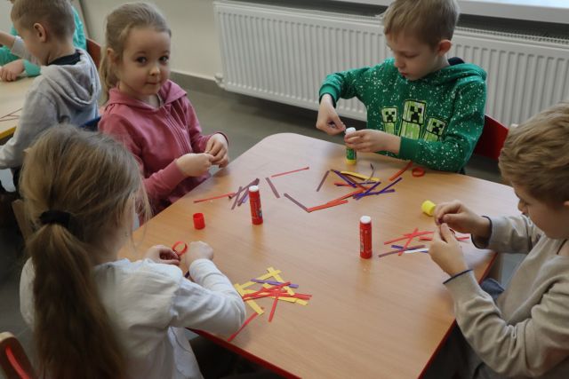 dzieci siedzą przy brązowym stole i wykonują z kawałków papieru łąńcuch na choinkę