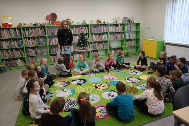 dzieci siedzą na zielonym dywanie, przed nimi stoi kobieta W CZARNEJ BLUZCE Z KSIĄŻKĄ W DŁONIACH, w tle regały z książkami