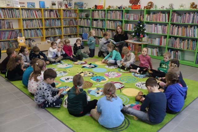 dzieci i kobieta czytająca książkę siedzą na zielonym dywanie, w tle regały z książkami