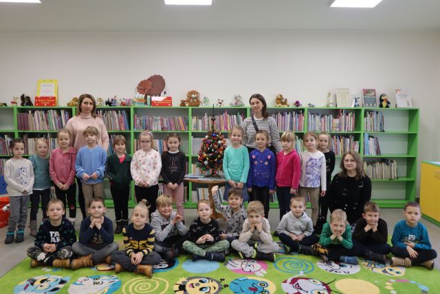 dzieci siedzą na zielonym dywanie, za nimi stoją dzieci, w tle regały z książkami