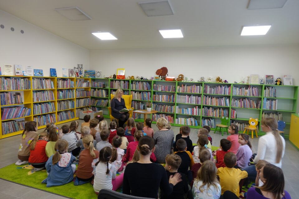 dzieci siedzą na zielonym dywanie, przed nimi siedzi na żółtym fotelu kobieta w siwej sukience i czyta książkę, w tle regały z książkami